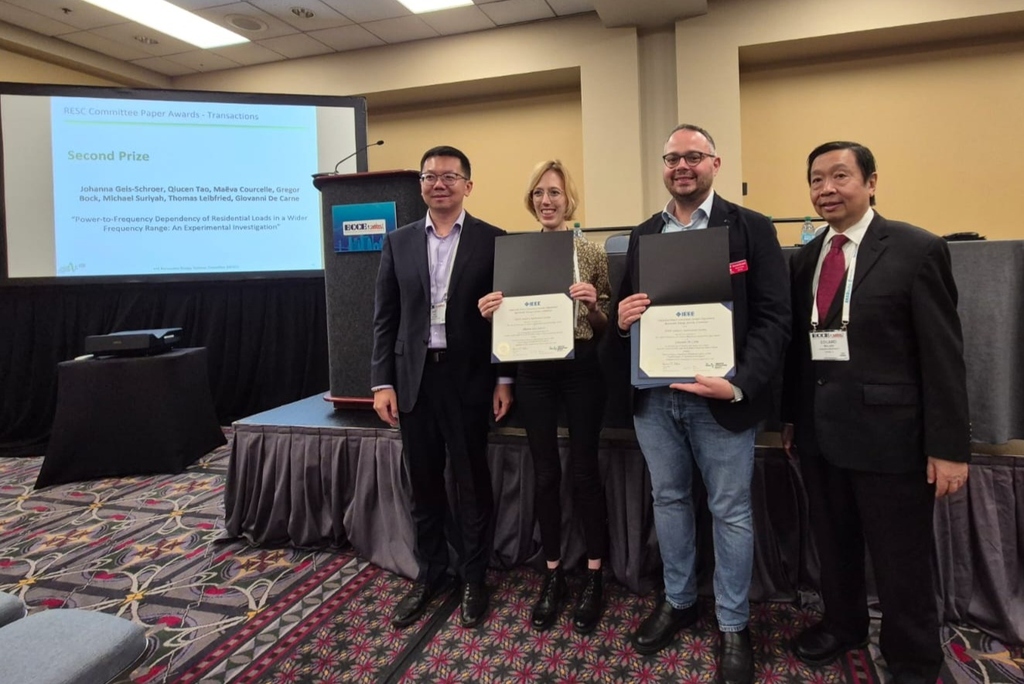 Ke Ma (Chair), Johanna Geis-Schroer (IEH), Giovanni de Carne (ITEP) und Eduard Muljadi (Vice Chair) stehen mit Urkunden vor einer Bühne bei einer wissenschaftlichen Preisverleihung.